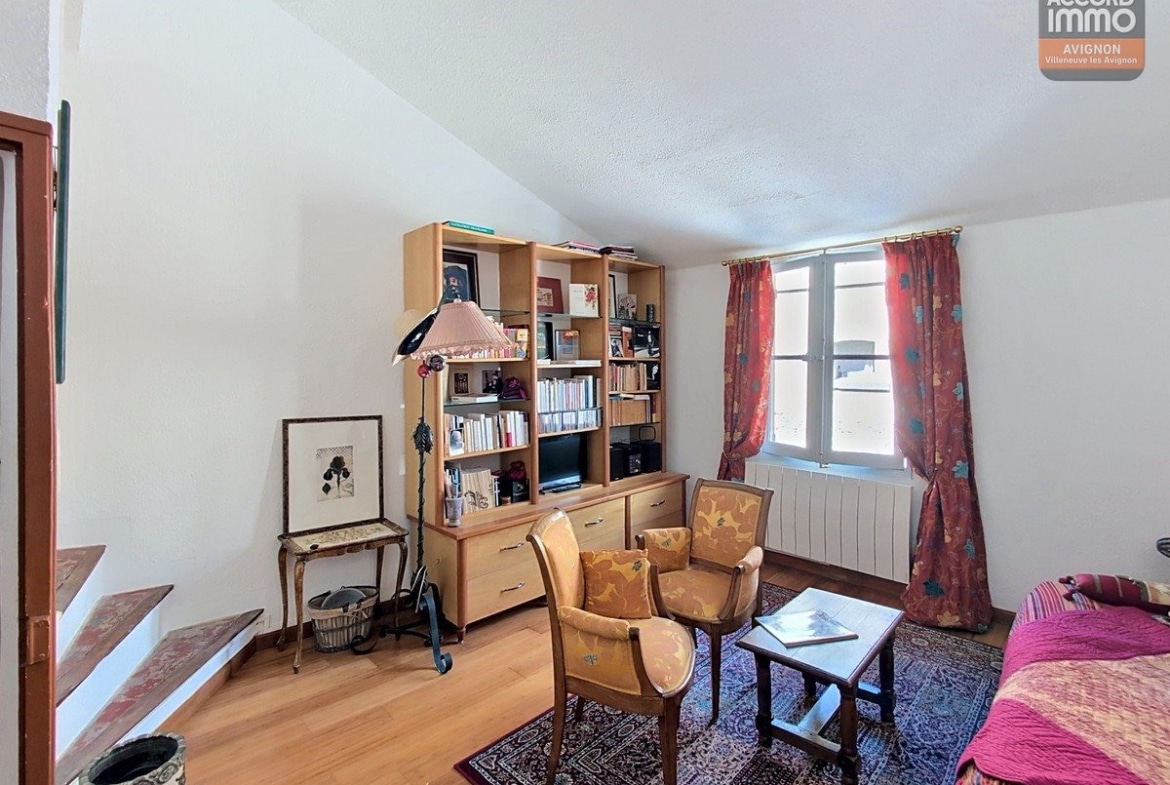 Pièce de vie mansardée avec bibliothèque modulable en bois clair et commode assortie sous la fenêtre. Fauteuil tapissé jaune, canapé rose, tapis oriental et escalier en pierre apparente menant à l'étage inférieur.