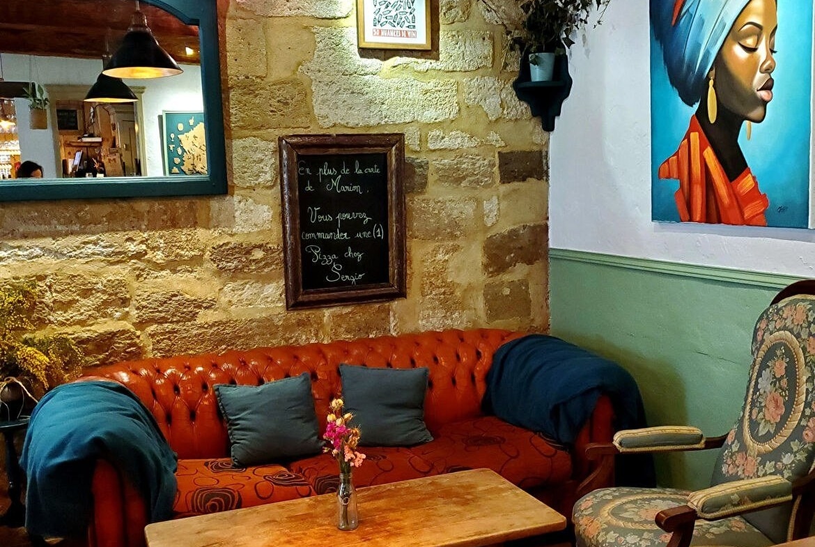 Espace lounge avec canapé Chesterfield orange, coussins bleus et table basse en bois sur parquet. Mur en pierre apparente orné d'un tableau portrait, d'un miroir vert et d'une ardoise menu manuscrite.