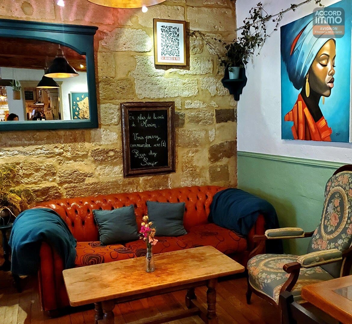 Espace lounge avec canapé Chesterfield orange, coussins bleus et table basse en bois sur parquet. Mur en pierre apparente orné d'un tableau portrait, d'un miroir vert et d'une ardoise menu manuscrite.