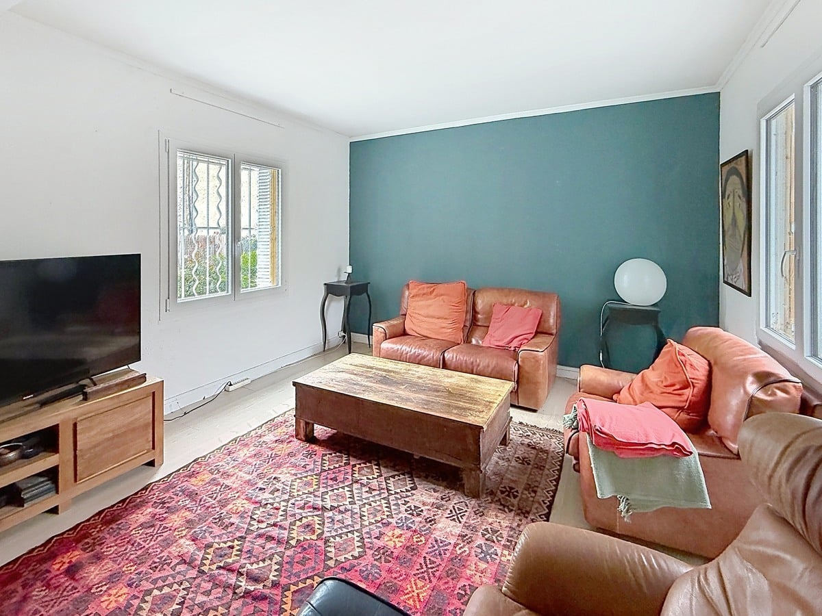 Salon avec trois fauteuils en cuir marron disposés autour d'une table basse en bois massif. Mur d'accent bleu canard, tapis kilim rouge à motifs géométriques et parquet clair.