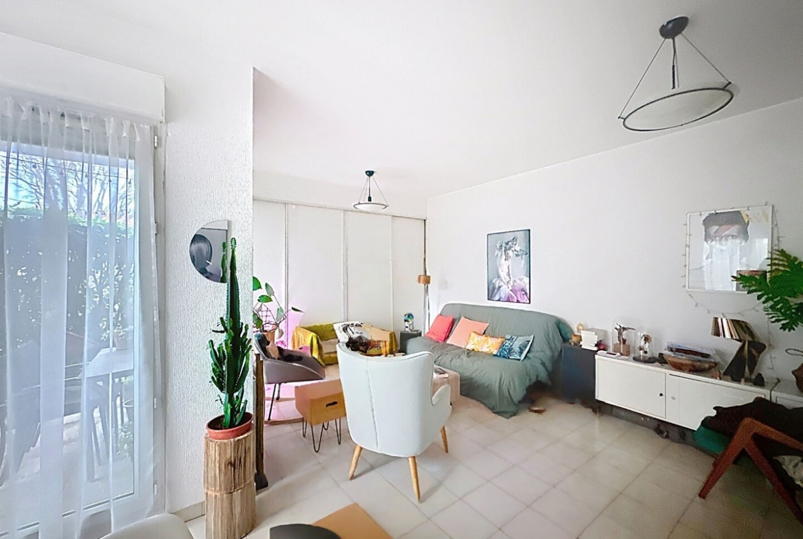 Pièce de vie avec carrelage beige au sol, canapé gris avec coussins colorés, fauteuil blanc à pieds bois et cactus en pot sur socle en bois. Baie vitrée avec voilage blanc donnant sur verdure, meuble TV blanc bas, suspensions circulaires noires au plafond et cadres décoratifs aux murs.