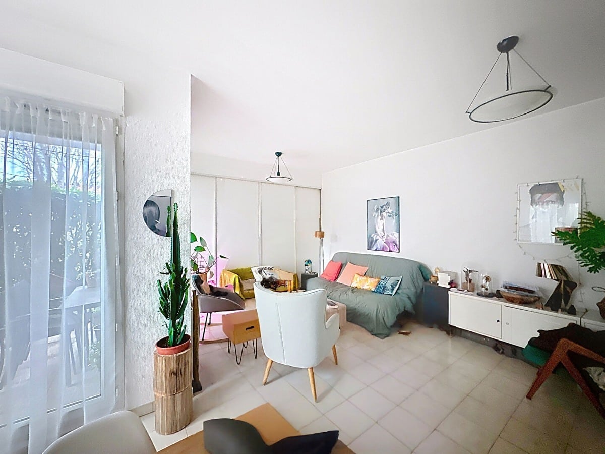 Pièce de vie avec carrelage beige au sol, canapé gris avec coussins colorés, fauteuil blanc à pieds bois et cactus en pot sur socle en bois. Baie vitrée avec voilage blanc donnant sur verdure, meuble TV blanc bas, suspensions circulaires noires au plafond et cadres décoratifs aux murs.