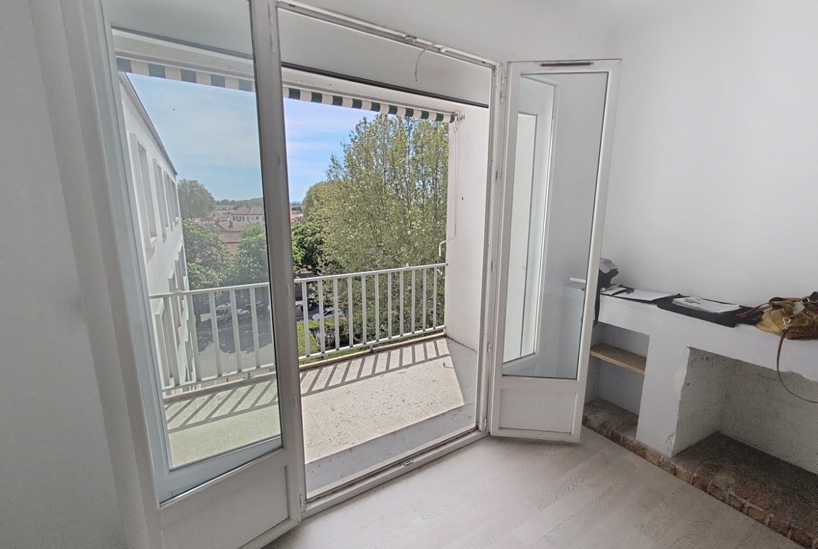 Balcon avec garde-corps à barreaux blancs et store banne rayé, vue sur parking arboré et toits de la ville. Ciel bleu avec quelques nuages, façade crépi blanc visible.