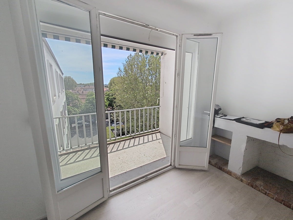 Balcon avec garde-corps à barreaux blancs et store banne rayé, vue sur parking arboré et toits de la ville. Ciel bleu avec quelques nuages, façade crépi blanc visible.