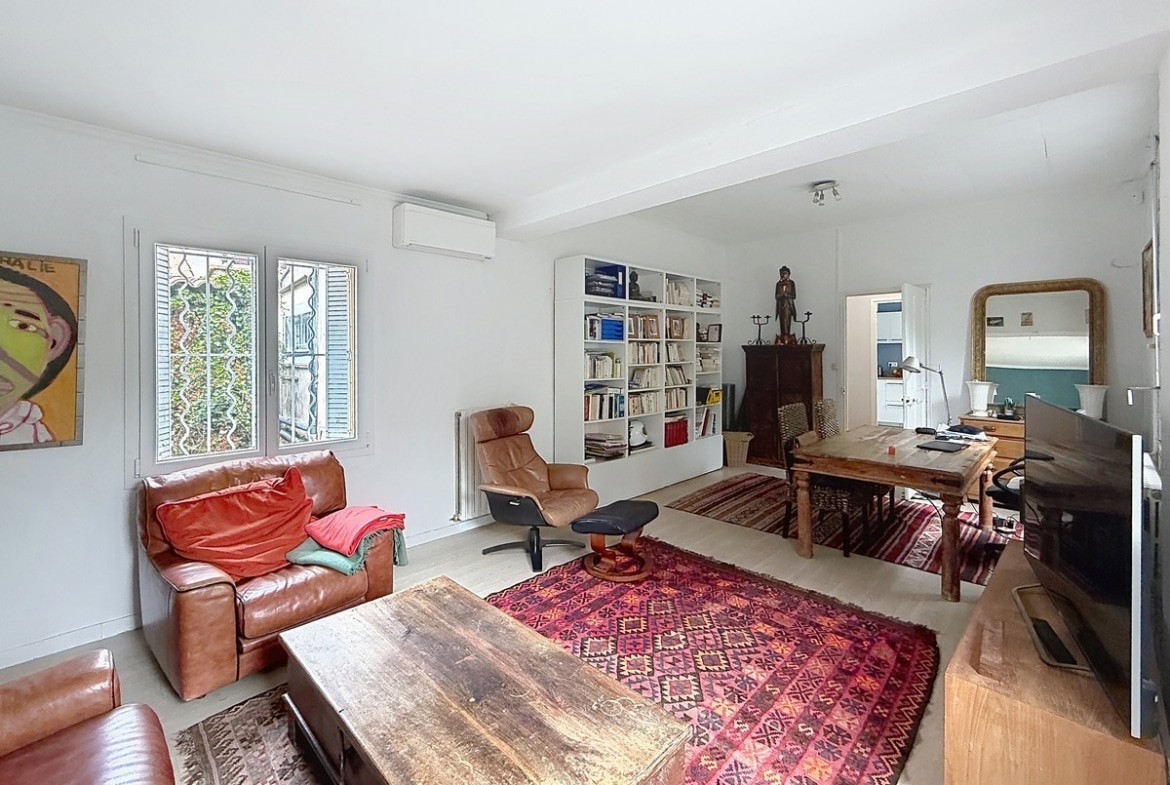 Pièce de vie ouverte avec canapés en cuir marron, fauteuil relaxant et bibliothèque blanche intégrée. Bureau en bois ancien, climatisation murale et miroir doré au mur.