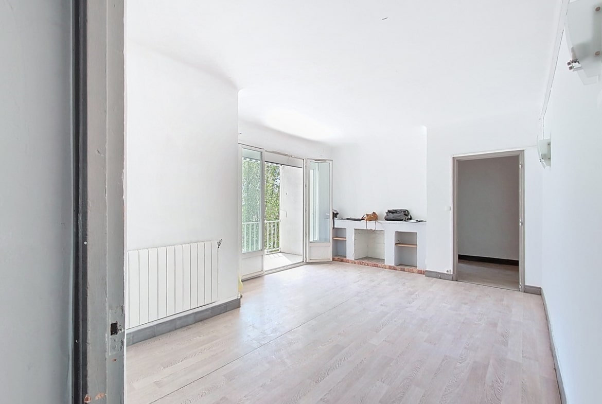 Grand séjour vide avec parquet clair, cheminée décorative en brique et étagères encastrées. Porte-fenêtre ouverte sur balcon, radiateur blanc sous fenêtre, murs blancs.