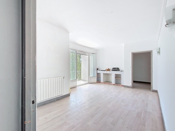 Grand séjour vide avec parquet clair, cheminée décorative en brique et étagères encastrées. Porte-fenêtre ouverte sur balcon, radiateur blanc sous fenêtre, murs blancs.