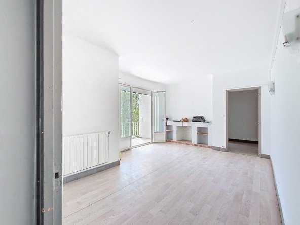Grand séjour vide avec parquet clair, cheminée décorative en brique et étagères encastrées. Porte-fenêtre ouverte sur balcon, radiateur blanc sous fenêtre, murs blancs.