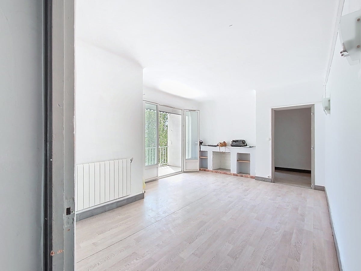 Grand séjour vide avec parquet clair, cheminée décorative en brique et étagères encastrées. Porte-fenêtre ouverte sur balcon, radiateur blanc sous fenêtre, murs blancs.