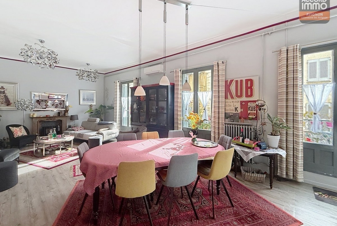 Vaste séjour avec cheminée ancienne en pierre sculptée, grande table ovale avec nappe rose entourée de chaises jaunes et bleues. Parquet clair, tapis persan rouge, enseigne vintage KUB rouge au mur. Quatre fenêtres avec rideaux à motifs et volets intérieurs gris, plafond haut avec moulures.