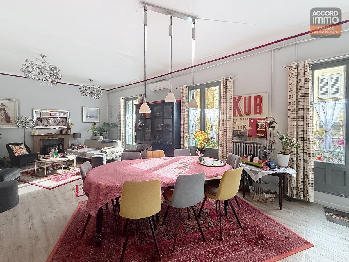 Vaste séjour avec cheminée ancienne en pierre sculptée, grande table ovale avec nappe rose entourée de chaises jaunes et bleues. Parquet clair, tapis persan rouge, enseigne vintage KUB rouge au mur. Quatre fenêtres avec rideaux à motifs et volets intérieurs gris, plafond haut avec moulures.