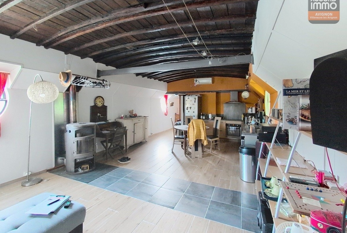 Espace de vie ouvert avec plafond voûté en bois sombre et poutres apparentes, poêle à bois ancien sur la gauche avec conduit métallique. Cuisine ouverte avec murs orangés, table à manger, sol en carrelage gris et parquet, climatisation murale visible.