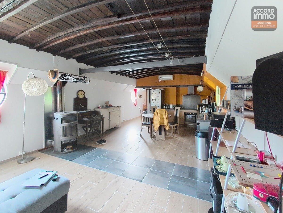 Espace de vie ouvert avec plafond voûté en bois sombre et poutres apparentes, poêle à bois ancien sur la gauche avec conduit métallique. Cuisine ouverte avec murs orangés, table à manger, sol en carrelage gris et parquet, climatisation murale visible.