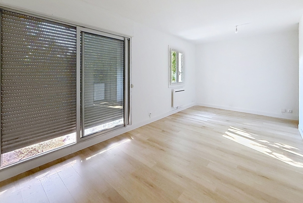 Pièce principale avec grande baie vitrée coulissante et fenêtre latérale apportant lumière naturelle. Sol en parquet clair, radiateur électrique blanc sous la fenêtre, murs peints en blanc.