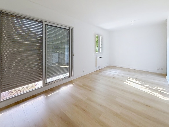 Pièce principale avec grande baie vitrée coulissante et fenêtre latérale apportant lumière naturelle. Sol en parquet clair, radiateur électrique blanc sous la fenêtre, murs peints en blanc.