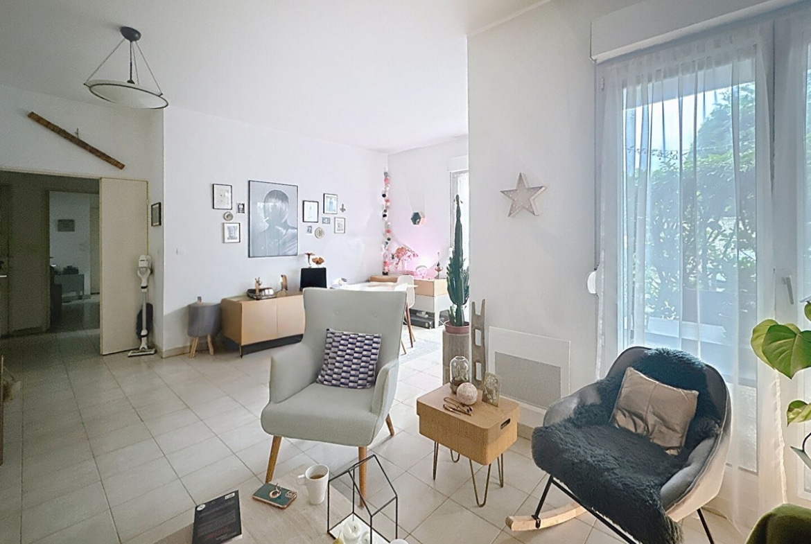 Espace de vie ouvert avec coin bureau au fond près de la fenêtre, fauteuil à bascule gris avec plaid bleu et fauteuil blanc. Table basse en bois avec objets décoratifs, carrelage clair au sol, étoile décorative blanche au mur et guirlande lumineuse colorée.