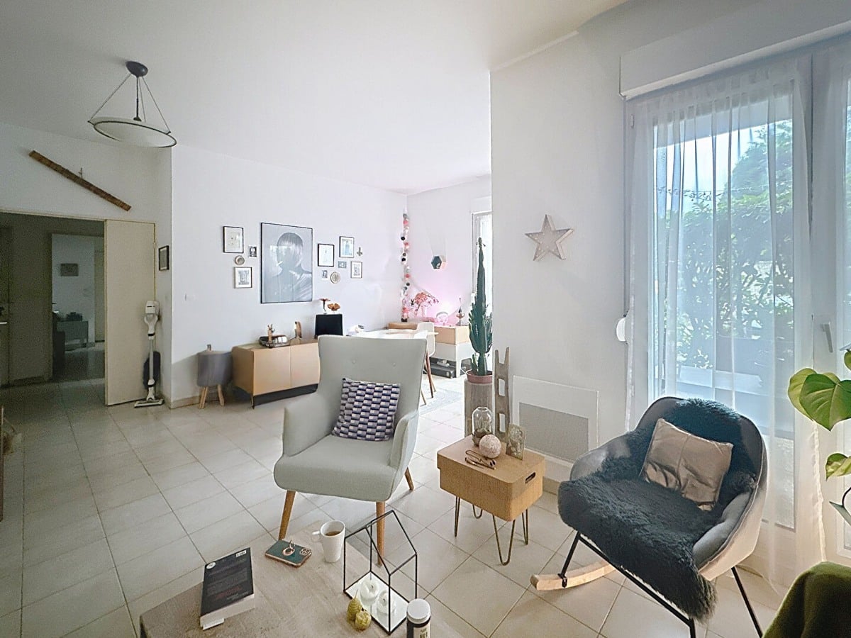 Espace de vie ouvert avec coin bureau au fond près de la fenêtre, fauteuil à bascule gris avec plaid bleu et fauteuil blanc. Table basse en bois avec objets décoratifs, carrelage clair au sol, étoile décorative blanche au mur et guirlande lumineuse colorée.