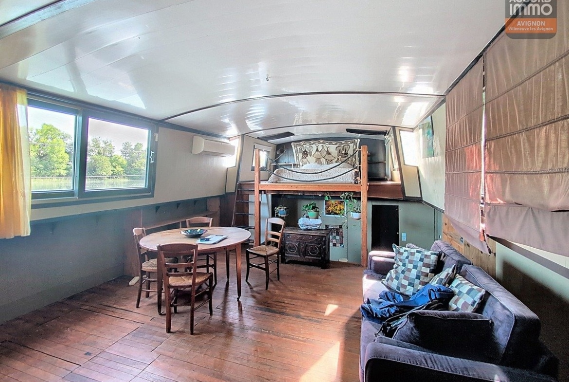 Intérieur de péniche avec plafond arrondi blanc, lit en mezzanine sur structure bois, canapé bleu foncé et table ronde avec chaises paillées. Parquet ancien au sol, fenêtres rectangulaires donnant sur l'extérieur verdoyant, tapisserie décorative au mur.
