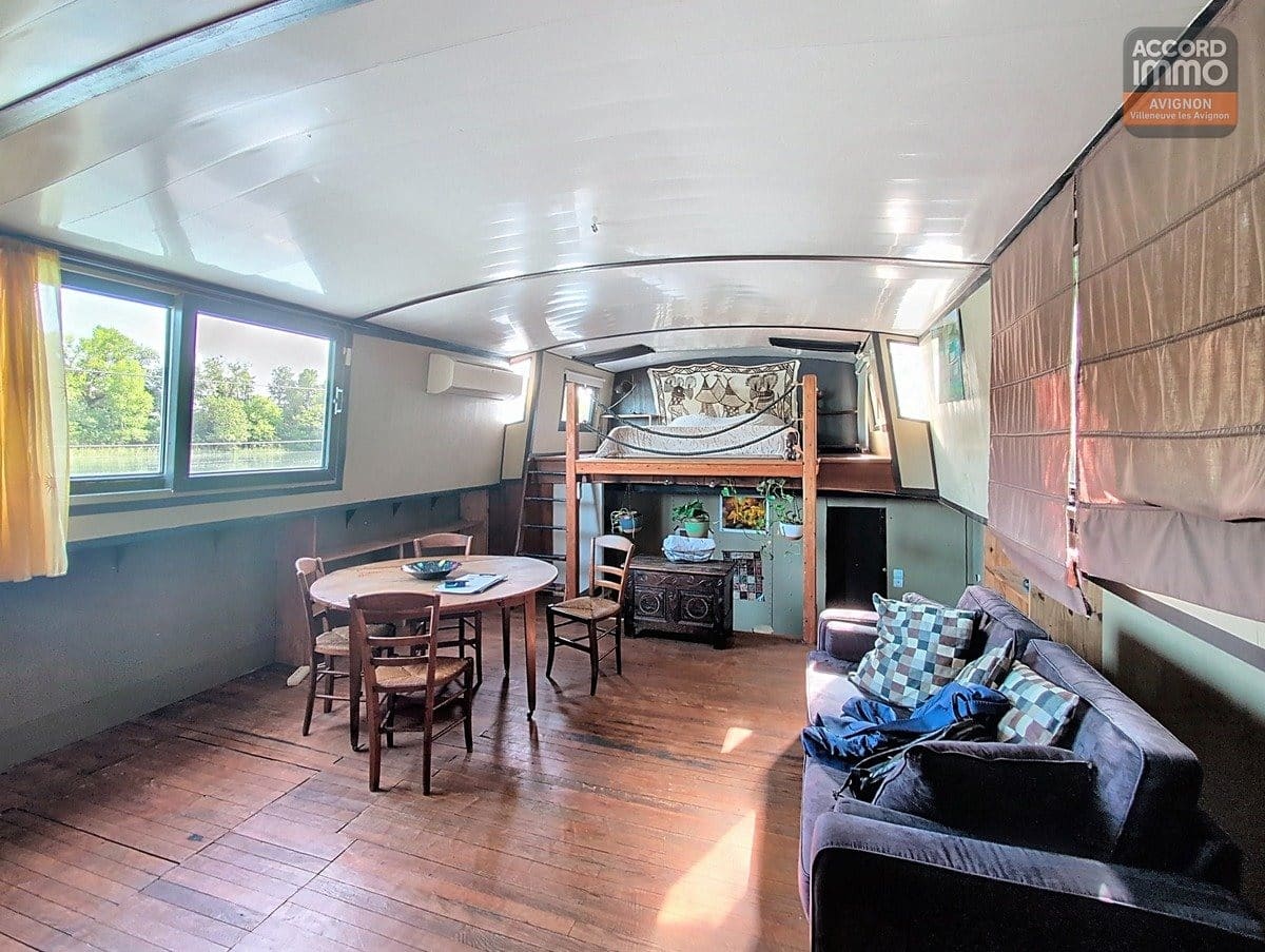 Intérieur de péniche avec plafond arrondi blanc, lit en mezzanine sur structure bois, canapé bleu foncé et table ronde avec chaises paillées. Parquet ancien au sol, fenêtres rectangulaires donnant sur l'extérieur verdoyant, tapisserie décorative au mur.