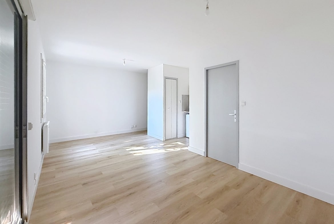 Pièce de vie avec sol en parquet stratifié clair et murs blancs. Baie vitrée coulissante avec volet roulant à demi fermé, deux portes grises menant vers cuisine et salle d'eau.