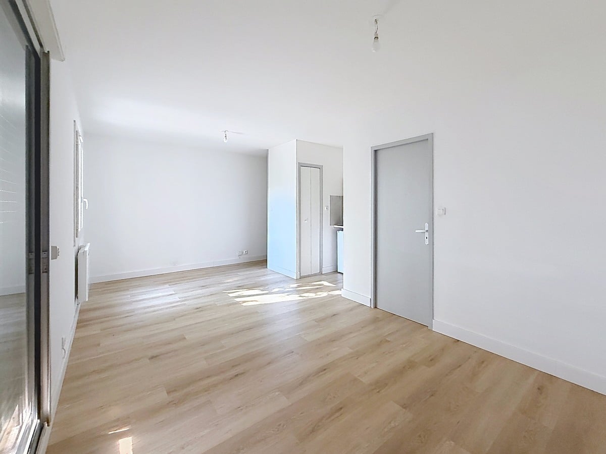 Pièce de vie avec sol en parquet stratifié clair et murs blancs. Baie vitrée coulissante avec volet roulant à demi fermé, deux portes grises menant vers cuisine et salle d'eau.