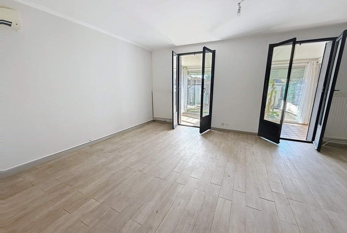 Pièce de vie vide avec sol imitation parquet clair et murs blancs. Deux portes-fenêtres à cadre noir aluminium ouvrent sur un espace extérieur, radiateur blanc sous fenêtre et climatiseur mural visible à gauche.
