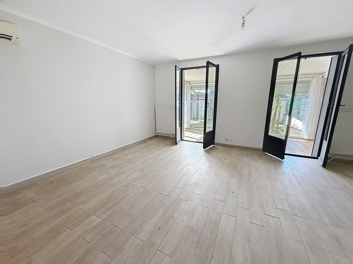 Pièce de vie vide avec sol imitation parquet clair et murs blancs. Deux portes-fenêtres à cadre noir aluminium ouvrent sur un espace extérieur, radiateur blanc sous fenêtre et climatiseur mural visible à gauche.