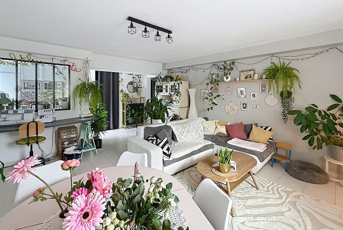 Pièce de vie ouverte avec canapé d'angle blanc et gris, table basse en bois scandinave et tapis à motifs ondulés. Table à manger ronde blanche avec chaises, verrière noire vers cuisine, nombreuses plantes suspendues et décoration murale bohème.