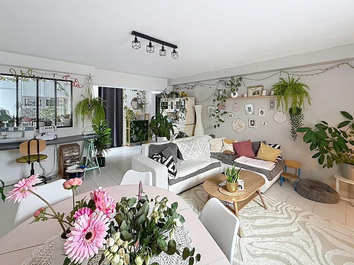 Pièce de vie ouverte avec canapé d'angle blanc et gris, table basse en bois scandinave et tapis à motifs ondulés. Table à manger ronde blanche avec chaises, verrière noire vers cuisine, nombreuses plantes suspendues et décoration murale bohème.