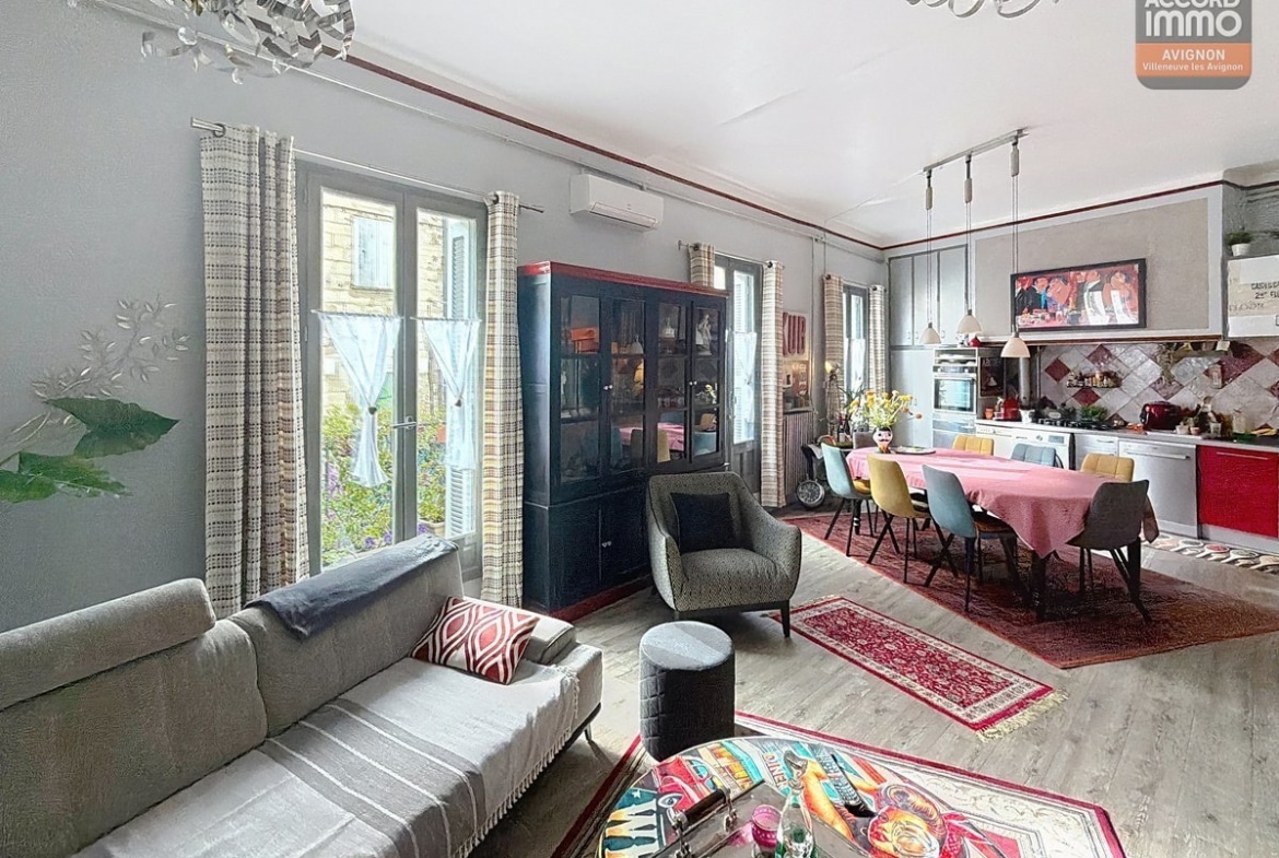 Grand séjour avec canapé gris, fauteuils assortis et table basse colorée, vaisselier noir vitré. Cuisine ouverte avec façades rouges et grises, table ovale avec nappe rose et chaises multicolores jaunes et bleues. Parquet clair, tapis orientaux rouges, hauts plafonds avec lustre design et climatisation murale.