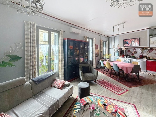 Grand séjour avec canapé gris, fauteuils assortis et table basse colorée, vaisselier noir vitré. Cuisine ouverte avec façades rouges et grises, table ovale avec nappe rose et chaises multicolores jaunes et bleues. Parquet clair, tapis orientaux rouges, hauts plafonds avec lustre design et climatisation murale.