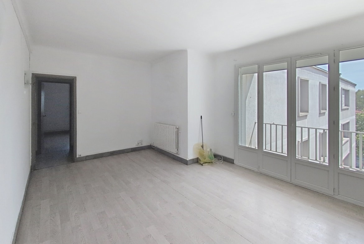 Séjour vide aux murs blancs avec parquet stratifié gris clair et radiateur mural. Grandes portes-fenêtres à petits carreaux donnant sur un balcon avec vue sur immeuble voisin.