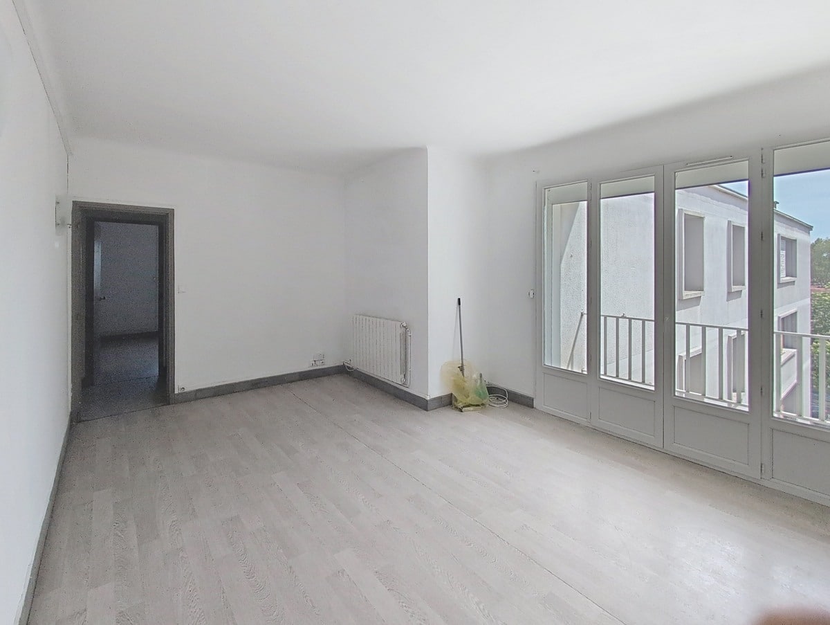 Séjour vide aux murs blancs avec parquet stratifié gris clair et radiateur mural. Grandes portes-fenêtres à petits carreaux donnant sur un balcon avec vue sur immeuble voisin.