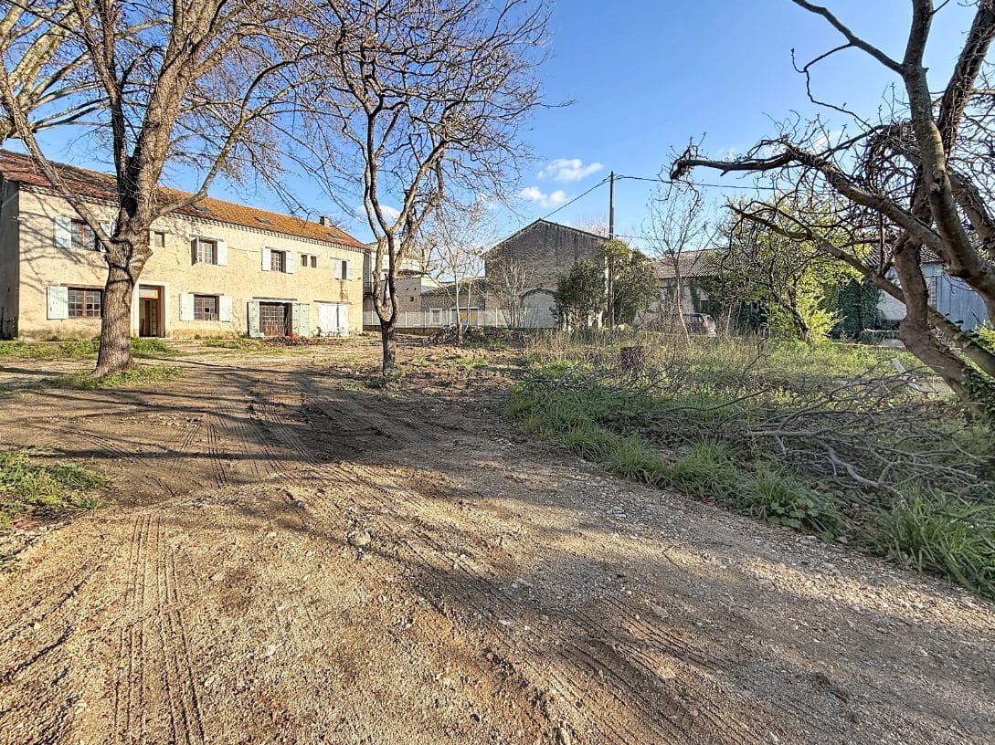 Vaste terrain en terre avec traces de passage de véhicules, bordé de grands arbres caducs sans feuilles et branches coupées au sol. Bâtiment principal en pierre enduite visible en arrière-plan avec dépendances en pierre et hangar, ciel bleu dégagé.