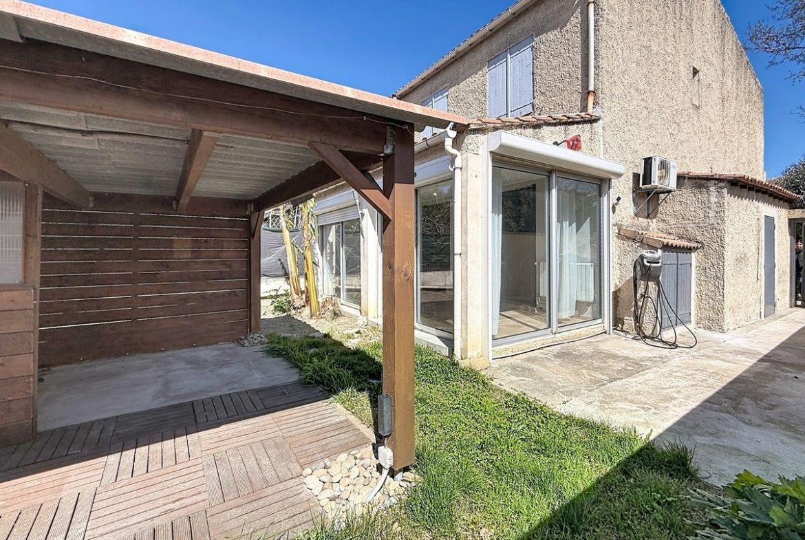 Extérieur avec terrasse en lames de bois composite et pergola couverte en bois brun. Maison mitoyenne à deux niveaux avec crépi beige, véranda vitrée aluminium et petit espace gazonné avec allée en pierre.