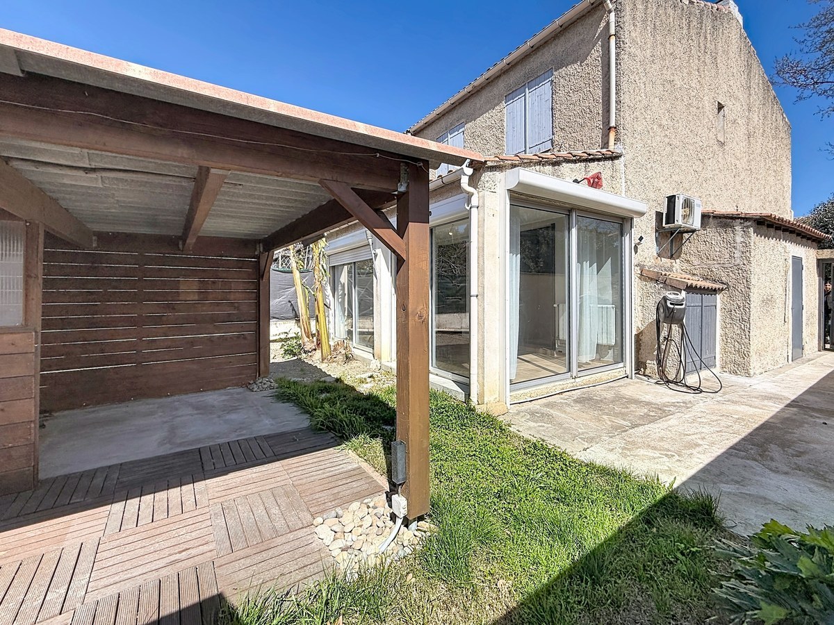 Extérieur avec terrasse en lames de bois composite et pergola couverte en bois brun. Maison mitoyenne à deux niveaux avec crépi beige, véranda vitrée aluminium et petit espace gazonné avec allée en pierre.