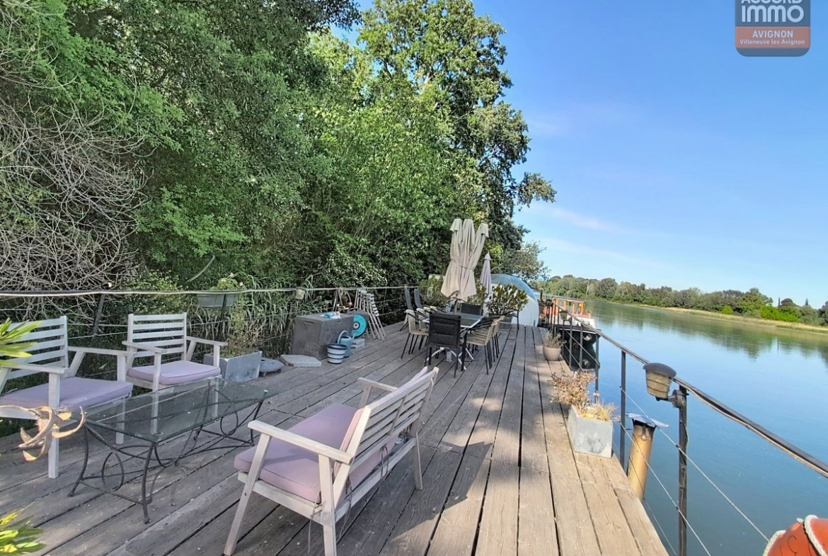 Grande terrasse en bois avec mobilier de jardin blanc et gris, parasol fermé et plantes en pots. Vue dégagée sur le Rhône et la végétation luxuriante de l'île de la Barthelasse, garde-corps en câbles métalliques, ciel bleu avec quelques nuages.