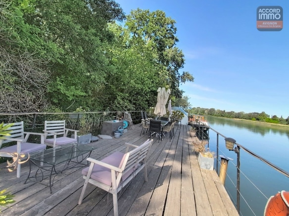 Grande terrasse en bois avec mobilier de jardin blanc et gris, parasol fermé et plantes en pots. Vue dégagée sur le Rhône et la végétation luxuriante de l'île de la Barthelasse, garde-corps en câbles métalliques, ciel bleu avec quelques nuages.
