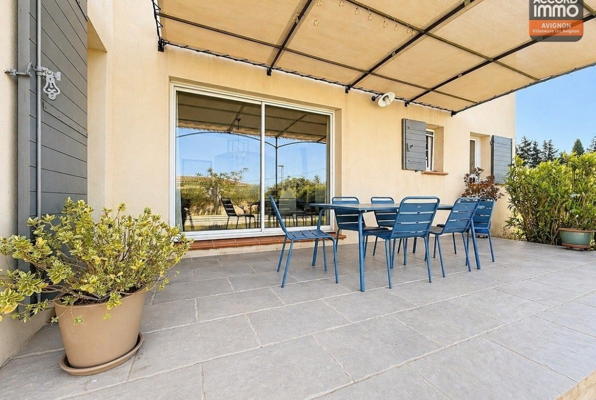 Terrasse dallée en pierre grise avec pergola métallique et toile tendue. Table et chaises bleu foncé pour six personnes. Vue sur le jardin avec piscine et oliviers en arrière-plan.