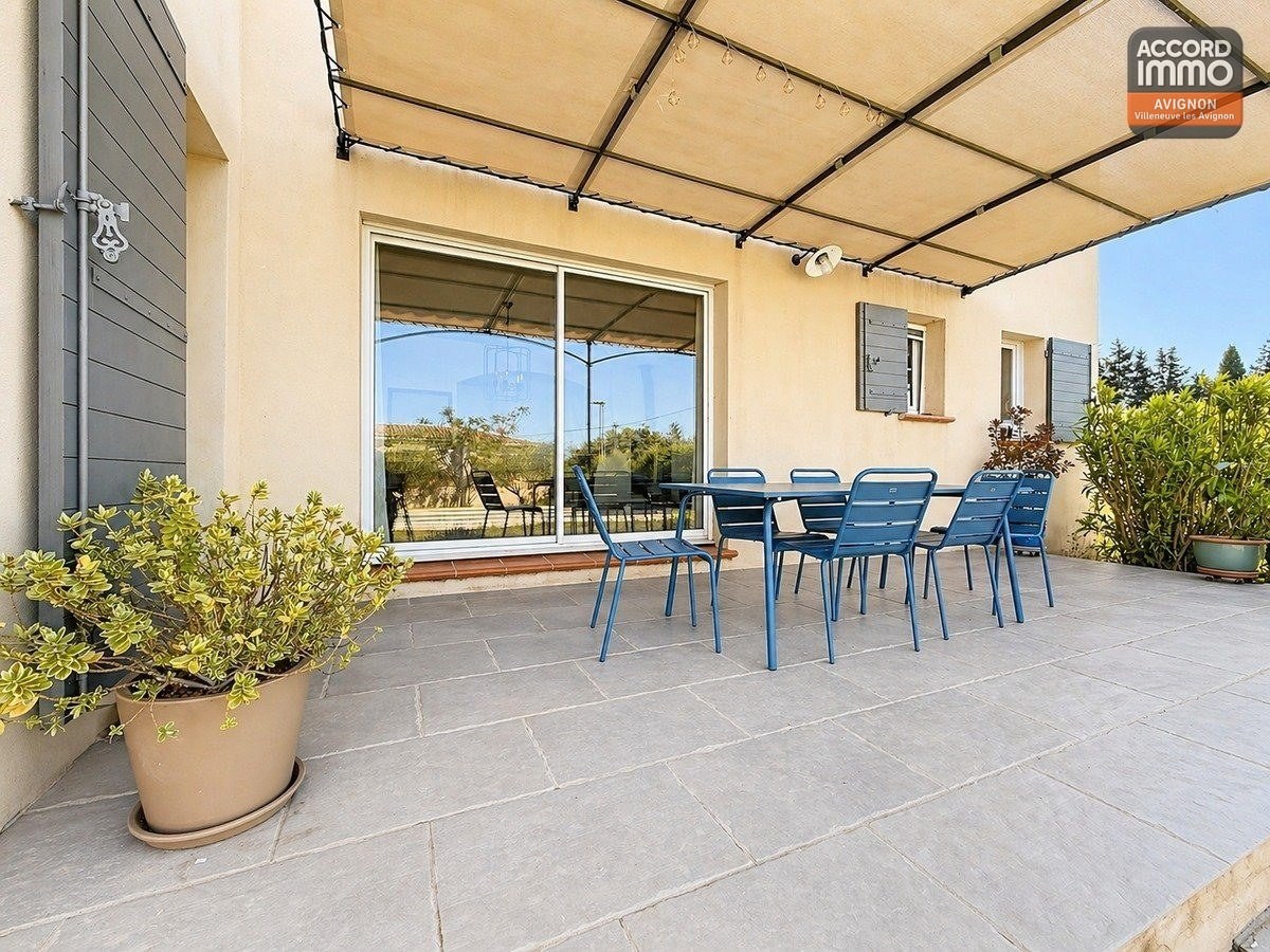 Terrasse dallée en pierre grise avec pergola métallique et toile tendue. Table et chaises bleu foncé pour six personnes. Vue sur le jardin avec piscine et oliviers en arrière-plan.