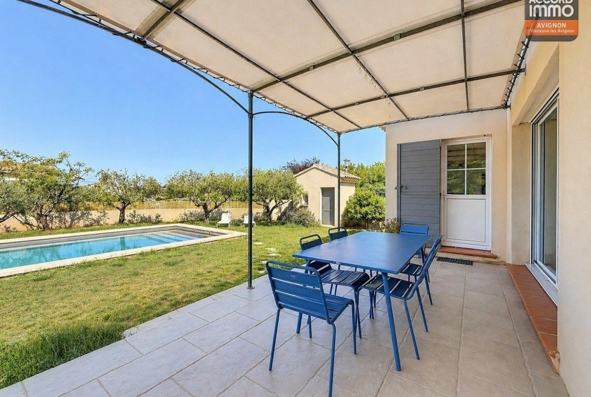 Terrasse couverte par une pergola avec toile blanche et structure métallique. Salon de jardin bleu sur dalles grises. Végétation méditerranéenne en pot et vue sur la piscine et le cabanon.