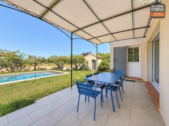 Terrasse couverte par une pergola avec toile blanche et structure métallique. Salon de jardin bleu sur dalles grises. Végétation méditerranéenne en pot et vue sur la piscine et le cabanon.