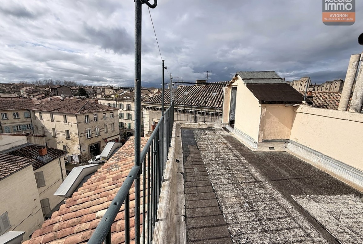 Terrasse étroite sur toiture avec garde-corps métallique noir, vue panoramique sur toits en tuiles provençales et bâtiments anciens du centre historique. Ciel nuageux, antenne et fils électriques visibles, cheminée blanche et lucarne en arrière-plan.