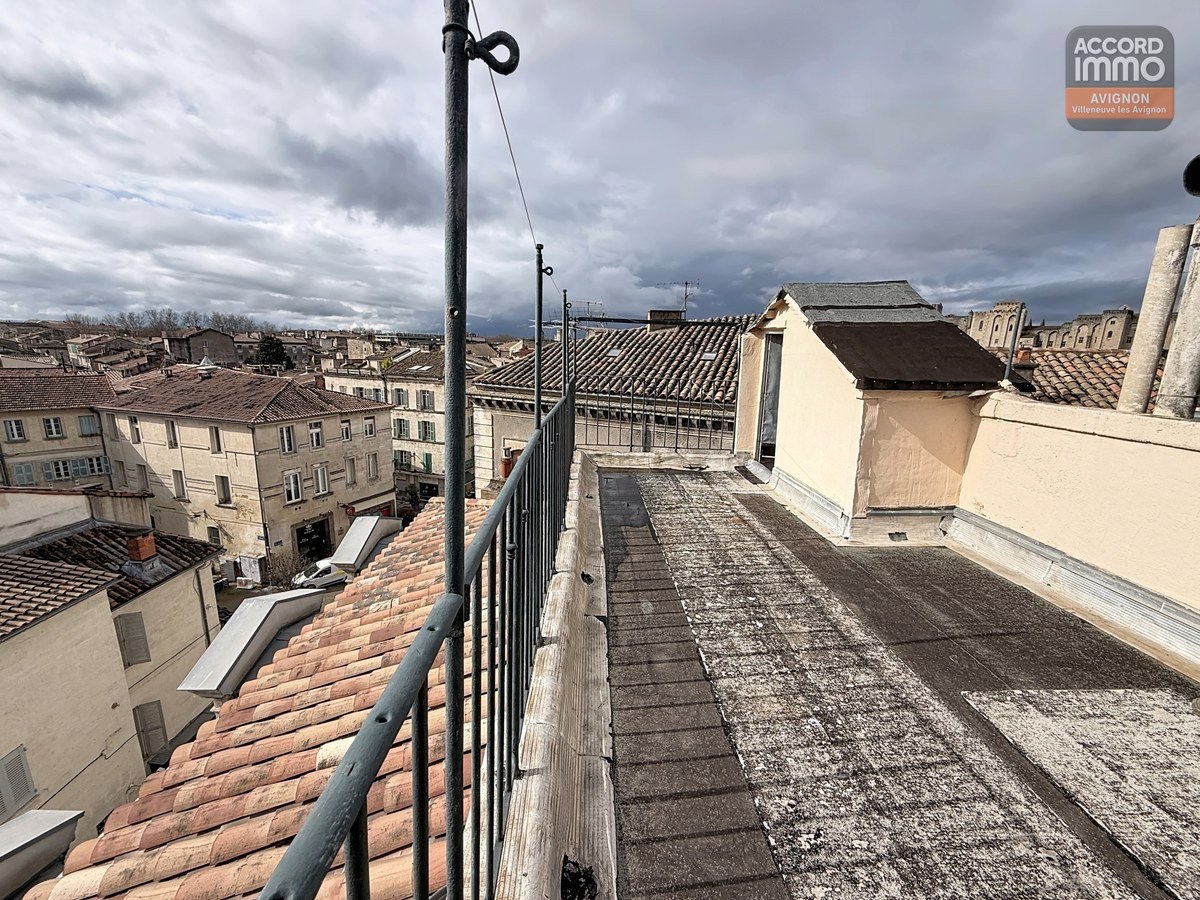 Terrasse étroite sur toiture avec garde-corps métallique noir, vue panoramique sur toits en tuiles provençales et bâtiments anciens du centre historique. Ciel nuageux, antenne et fils électriques visibles, cheminée blanche et lucarne en arrière-plan.