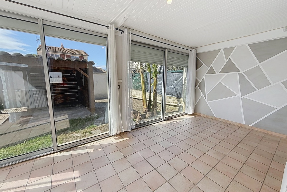 Véranda carrelée en terre cuite avec plafond en lambris blanc. Grandes baies vitrées aluminium avec volets roulants donnant sur jardin, mur décoré de motifs géométriques gris et blancs.