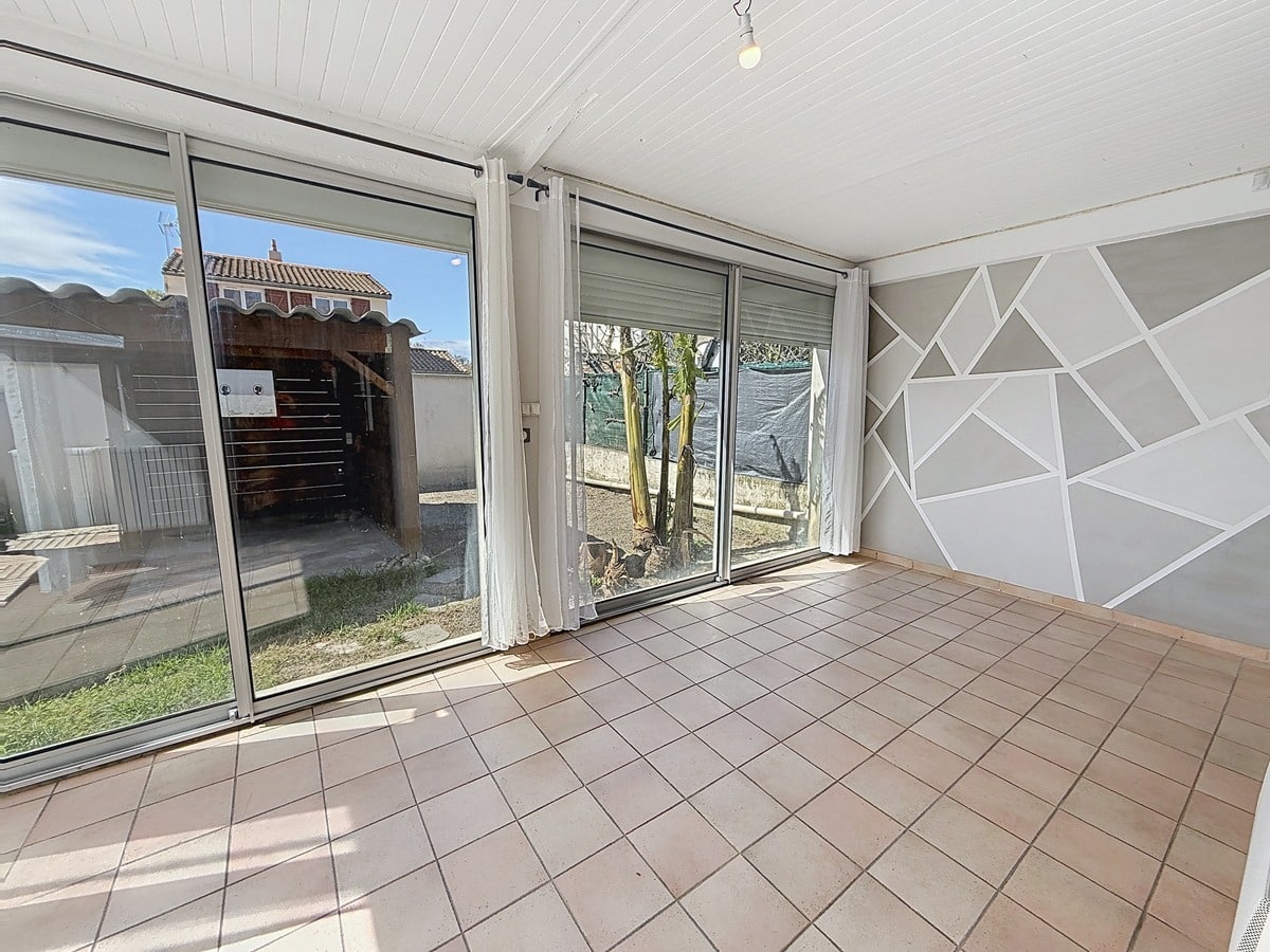 Véranda carrelée en terre cuite avec plafond en lambris blanc. Grandes baies vitrées aluminium avec volets roulants donnant sur jardin, mur décoré de motifs géométriques gris et blancs.