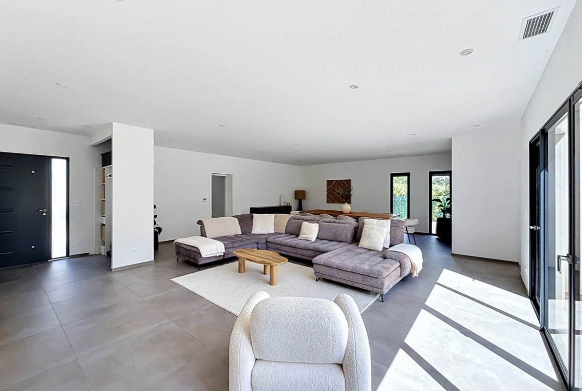 Vaste espace de vie avec sol en carrelage grand format gris et murs blancs. Canapé d'angle gris, fauteuil bouclé blanc et table basse en bois naturel avec lumière naturelle par les baies vitrées.
