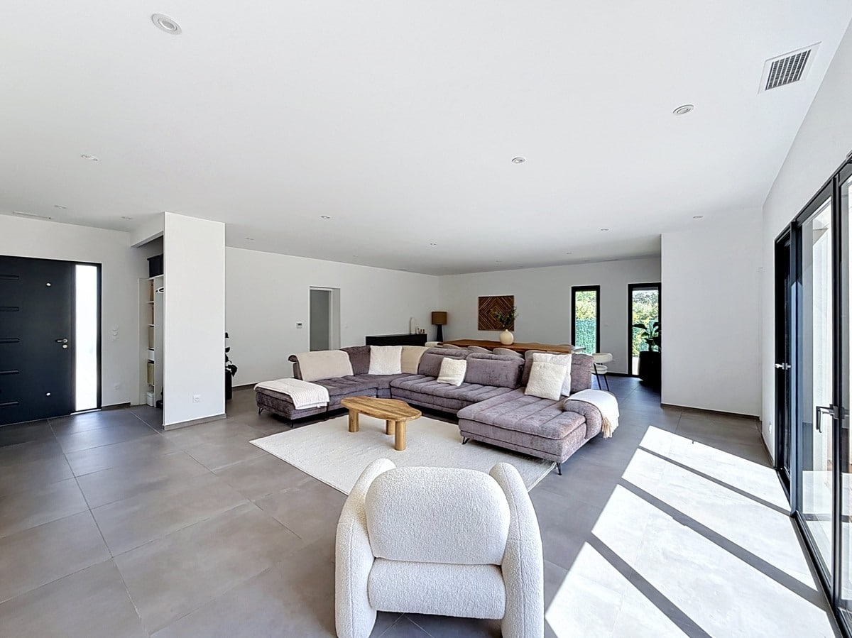 Vaste espace de vie avec sol en carrelage grand format gris et murs blancs. Canapé d'angle gris, fauteuil bouclé blanc et table basse en bois naturel avec lumière naturelle par les baies vitrées.