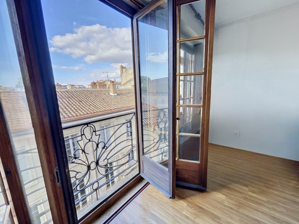 Portes-fenêtres en bois foncé ouvertes sur un balcon avec garde-corps en fer forgé décoratif. Vue dégagée sur toits en tuiles et façades anciennes, ciel bleu avec nuages blancs, parquet clair dans la pièce.