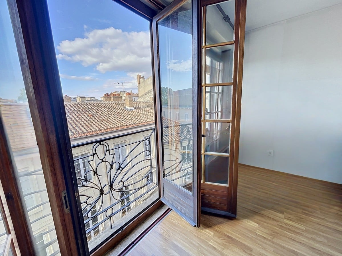 Portes-fenêtres en bois foncé ouvertes sur un balcon avec garde-corps en fer forgé décoratif. Vue dégagée sur toits en tuiles et façades anciennes, ciel bleu avec nuages blancs, parquet clair dans la pièce.